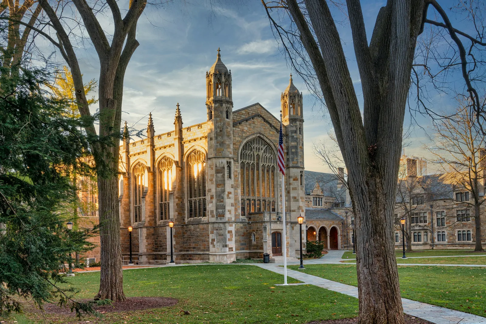 Evening sun lighting the Gothic windows of a stone campus hall in Ann Arbor, framed by bare autumn trees and lit lamp posts