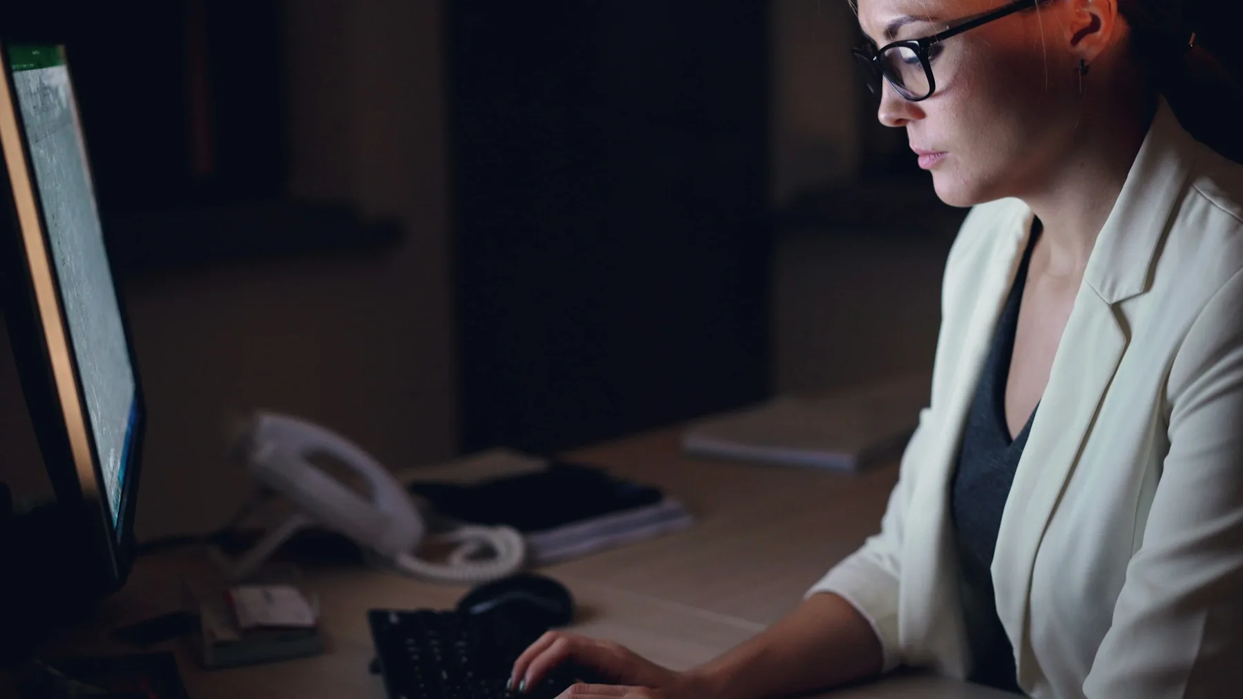 Woman in glasses focused on her computer at a desk in a dark office at night