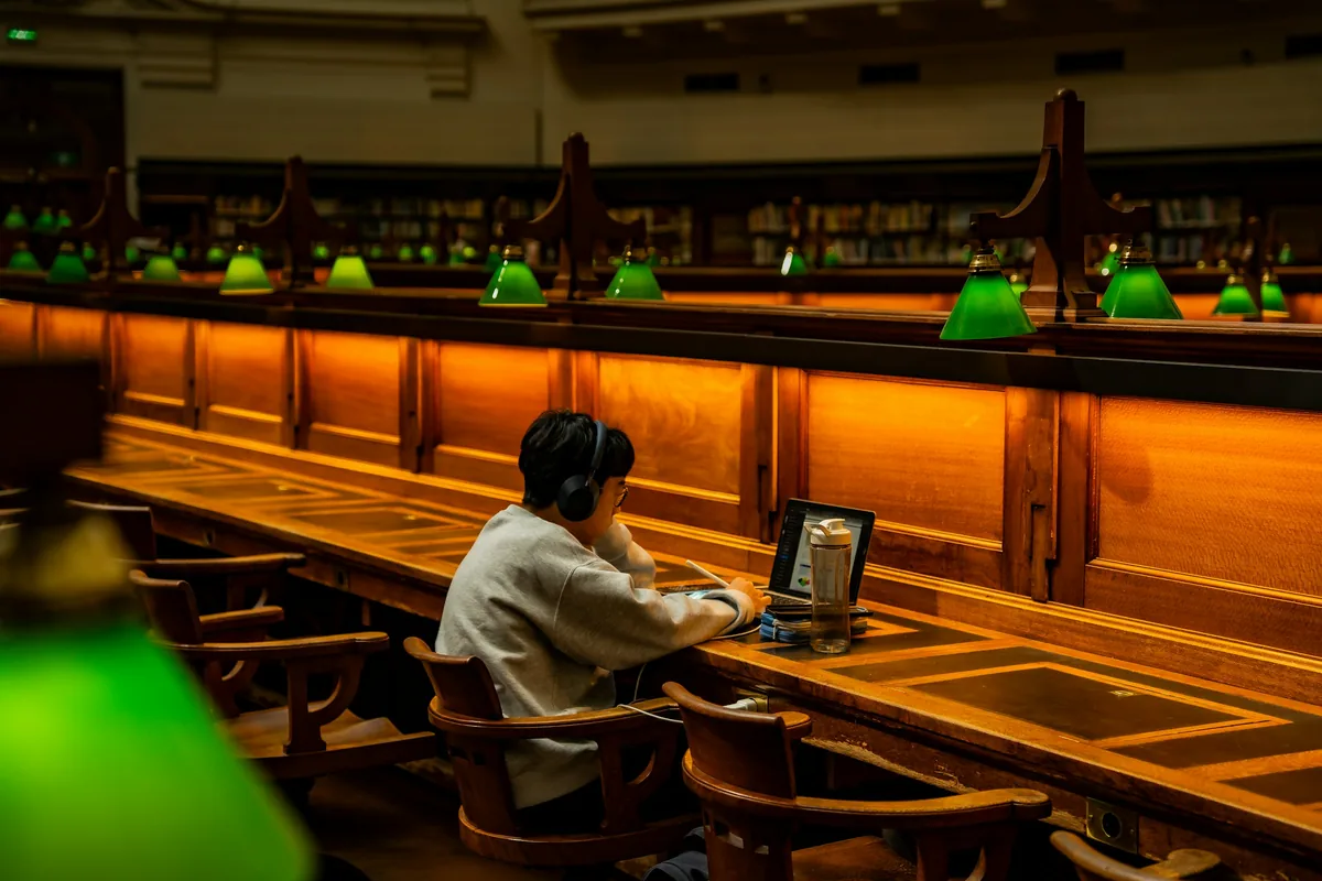 Student in headphones working on a laptop at a long wooden table in a library